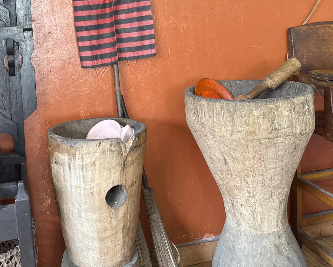 Rincón del comedor con un tambor de madera tallada, un pilón con su mano y una camisilla rayada colgada en la pared color terracota